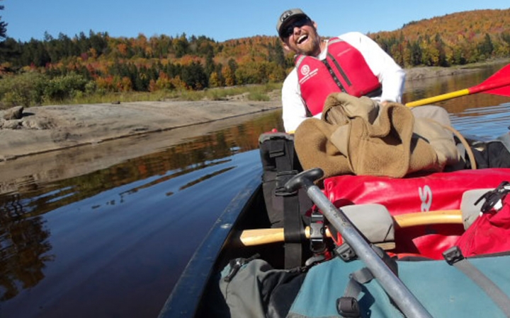 A person sitting in the back of a canoe smiles at the camera. There is fall foliage on the shore behind them.